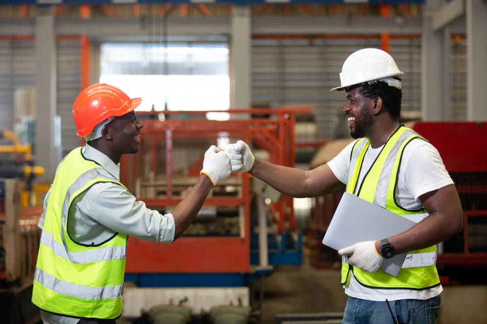 Two professionals wearing hard hats, gloves and safety vests in a workshop.